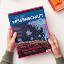 Auf einer weißen Fläche hält eine nicht näher bestimmbare Person mit zwei Händen und grauen Strickärmeln schräg das Magazin „BILDER DER WISSENSCHAFT“ mit pinkem Rand und blauem Titelbereich. Das Cover zeigt quallenähnliche, durchscheinende Organismen über einer violett-braunen Oberfläche und trägt die Texte „Unser Immunsystem“, „Im Fokus: Monde mit Tiefenmeeren: Verbergen sich dort exotische Lebensformen?“, „Bestäuber“ und „Smart Home“; oben rechts liegt teilweise ein orangefarbenes Objekt.