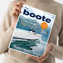 Weibliche Person im beigefarbenen Strickpullover hält ein schräg ausgerichtetes Magazin mit dem Titel „boote“. Das Cover zeigt ein weißes Motorboot auf blauem Wasser vor felsigen Bergen und einem Segelboot; lesbare Texte sind „Unter Strom!“, „Ruhe im Schiff“, „Boot-Zubehör?“, „Im Test“, „Reise“ und „Stern von Berlin“.