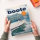 Schräg auf einem weißen Tisch gehaltenes Magazin „boote“ mit dunkelblauem Titel, blauem See, Berglandschaft und weißem Motorboot mit einer steuernden Person. Sichtbare Texte: „Unter Strom!“, „Ruhe im Schiff“ und „Stern von Berlin“; zwei Hände halten das Magazin, das Geschlecht ist nicht erkennbar.