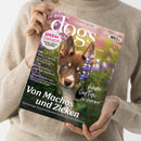 Person in beigefarbenem Strickpullover hält ein Magazin mit dem Titel „dogs – Vom Glück, mit Hunden zu leben“, Ausgabe 3/2025. Auf dem Cover ist ein braun-weißer Hundewelpe mit blauen Augen zwischen violett-blauen Blumen abgebildet. Sichtbare Texte: „Spezial Tierarzt: Kosten, Stress & Notfälle“, „Von Machos und Zicken“, „Perfekter Garten für Vierbeiner“, „Pro & Contra Tierschutz im Ausland“, „Checklist Europa – Urlaub mit Hund“.