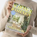 Hände einer Person in beigefarbenem Strickpullover halten das Wohnmagazin „SCHÖNER WOHNEN“ mit gelber Titelschrift und Unterzeile „Europas größtes Wohnmagazin“. Auf dem Cover ist ein heller Raum mit Holztisch, Stühlen, grünen Fensterläden und vielen Pflanzen abgebildet. Sichtbare Texte: „FERIEN-FLAIR“, „Sommerhäuser von Dänemark bis Marokko“, „Outdoor-Tipps“, „Schlaf-Guide für heiße Nächte“, „Komfortzone“, „An der Garden Route“.