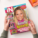 Titelseite der Zeitschrift „IN“ mit großem Porträt einer lächelnden blonden Frau auf pinkfarbenem Hintergrund, Hauptüberschrift „Beauty‑Beichte! So steht sie wirklich zu Botox & Co.“ in gelber und weißer Schrift; seitlich kleinere Bilder weiterer Personen mit Texten zu Themen wie Babyplänen, Klinikaufenthalt, Vorwürfen und Kriminalfall; Hände halten das Magazin über einem hellen Tisch mit orangefarbenem Objekt.