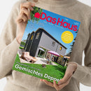 Helle, neutral gekleidete Person hält ein Magazin mit dem Titel „Das Haus“ vor hellem Hintergrund, Cover zeigt ein modernes, dunkelgraues Einfamilienhaus mit gelbem Fensterrahmen, Sonnenschirm und Personen auf einer Terrasse im Garten. Sichtbare Texte: „Das Haus – Bauen, Wohnen, Schöner Leben“, „Gemischtes Doppel“, „Klug geplant für zwei Familien“, „Tomaten – Anbautipps und besondere Sorten für Ihren Garten“.