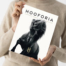 Person in beigem Strickpullover hält ein Magazin mit weißem Cover vertikal vor dem Oberkörper, darauf in schwarzer Schrift der Titel „HOOFORIA“ und darunter der Text „NACHDENKEN • BEWEGEN • VERSTEHEN • BEGEISTERN“. Zentral abgebildet ist ein schwarzes Pferd mit wehender Mähne auf hellem Hintergrund, unten mittig ein goldenes Monogramm „HC“ mit dem Zusatz „VOLUME 1“.