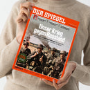 Person in beigem Strickpullover hält ein Exemplar des Magazins „Der Spiegel“, Ausgabe Nr. 26 / 19. 6. 2026, mit orangefarbenem Rand und Titel „Unser Krieg gegen Russland“. Das Titelbild zeigt vier Soldaten in dunklen Uniformen und Helmen mit Waffen auf einem Feld, im Hintergrund Rauch und Explosionen. Weitere sichtbare Textelemente: „Vor 85 Jahren überfiel NAZI-DEUTSCHLAND die Sowjetunion“ und ein grüner Kreis mit „Im Heft: Gratis-Zugang zur erweiterten MAZ-Karte“.