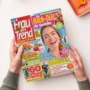 Titelblatt der Zeitschrift „Frau im Trend“ mit großem Foto einer lachenden Frau vor blauem Hintergrund. Mehrfarbige Schlagzeilen in Rosa, Gelb, Blau und Weiß, u. a. „Die Hack-Diät für Genießer“, „60 Pfund weg“, „Sofort 5 Jahre jünger aussehen“ und „Sparen – so einfach wie nie“. Abgebildet sind mehrere kleine Essensfotos, Textfelder und das rote Magazinlogo links oben. Zwei Hände halten die Zeitschrift über einem hellen Tisch.