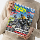 Person in beigefarbenem Pullover hält ein Magazin mit dem Titel „TOUREN FAHRER“ in großen gelben Buchstaben auf blauem Hintergrund. Das Cover zeigt drei Motorradfahrende in Schutzkleidung auf Landstraße, Überschriften u. a.: „NIE WIEDER KUPPELN?“, „SCHON GEFAHREN: APRILIA Tuareg Rally 660, HONDA GB 350 S“ und „BMW R 1300 RT“. Oben zusätzliche Themen zu Tourenkombis, Tankrucksäcken und Frühlingstests.