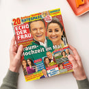 Titelblatt der Zeitschrift „Echo der Frau“ mit großem Porträt eines lachenden Mannes in grüner Jacke und einer lachenden Frau mit zurückgebundenem Haar vor rotem Hintergrund. Hauptüberschrift: „Traum-Hochzeit im Mai“. Weitere Textfelder und kleinere Porträtfotos von verschiedenen Personen sowie Schlagzeilen zu Themen wie Prominente, Liebe und royale Nachrichten in gelben, roten und blauen Kästen. Hände halten das Magazin über hellem Tisch.