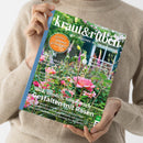 Person in beigem Strickpullover hält die Zeitschrift „kraut&rüben – Schöner. Wilder. Biogarten.“ (Ausgabe 06/2025) senkrecht in beiden Händen; auf dem Titelbild ein sommerlicher Garten mit rosafarbenen Rosen und grünen Stauden. Sichtbare Texte: „Großes Sommer-Gewinnspiel“, „Gestalten mit Rosen“, „Extra-Beilage: Mit Haustieren natürlich leben“, „Sauerkirschen – So finden Sie die richtige Sorte“, „Schneckenfeste Blütenstauden“.