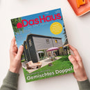 Titelbild der Zeitschrift „Das Haus“ mit blauem Schriftkopf und rotem Logo. Abgebildet ist ein modernes graues Doppelhaus mit großem Garten, Terrasse, Liege, Stühlen und Sonnenschirmen; zwei Personen sitzen im Außenbereich. Text auf dem Cover: „Gemischtes Doppel“, „Klug geplant für zwei Familien“, „Tomaten – Anbautipps und besondere Sorten für Ihren Garten“ sowie Hinweise zu Haushaltsgeräten und Bauinterview.