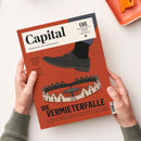 Rotes Capital-Magazin mit schwarzem Lederschuh und einer Illustration kreisförmig angeordneter Häuser auf dem Cover, von zwei Händen über weißer Fläche gehalten. Sichtbare Texte: „Capital“, „WIRTSCHAFT IST GESELLSCHAFT“, „CHINA-GEFAHR – Die große Warenflut kommt“, „AKTIVE FONDS“ und „DIE VERMIETERFALLE“; oben rechts liegt ein orangefarbenes Notizbuch mit Locher.