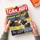 Auf weißer Fläche hält eine nicht näher erkennbare Person mit beiden Händen eine schräg liegende Zeitschrift „CAR&HIFI“ mit rot-gelbem Cover. Sichtbar sind Abbildungen von Verstärkern, Lautsprechern und Autoradios sowie die Texte „Sound mit DSP“, „Mini Endstufen“, „Edle Lautsprecher“, „Bass extrem“ und „Großbildschirm nachgerüstet“; oben rechts liegt teilweise ein orangefarbener Gegenstand.