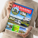 Weibliche Person im beigefarbenen Strickpullover hält ein schräg ausgerichtetes, farbiges Magazin mit dem Titel „CARAVANING – Europas großes Camping-Magazin“. Auf dem Cover sind mehrere weiße Wohnwagen, ein schwarzes Auto, Landschaftsfotos sowie die Texte „Neuheiten 2027“, „ADRIA ALPINA“, „FENDT APERO“, „TABBERT VIVALDI“, „Urlaub am Wasser“ und „Camping im Loiretal“ zu sehen.