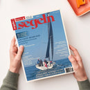 Titelblatt der Zeitschrift „segeln“ mit rotem Schriftzug auf blauem Hintergrund, im Mittelpunkt ein Segelboot mit schwarzen Segeln und mehreren Personen mit farbiger Segelkleidung auf dem Deck. Textausschnitte u. a.: „TOP YACHT AWARD 2025“, „NIEDERLANDE, NORDSEE, OSTSEE“, „GROSSER TÖRN MIT KLEINEM BOOT“. Das Magazin wird von zwei Händen auf hellem Tisch mit orangefarbenem Tablett gehalten.