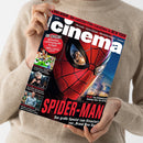 Weibliche Person im hellbeigen Strickpullover hält ein schräg ausgerichtetes Magazin mit dem Titel „in“ vor dem Oberkörper. Das Cover zeigt eine rote Spider-Man-Maske und schwarze Netzlinien; sichtbare Texte sind „EXKLUSIVE Die coolsten Film-Locations in NEW YORK“, „SPIDER-MAN“, „Das große Special zum Kinostart von ‚Brand New Day‘“ sowie „PLUS Tom Holland und Zendaya über den Dreh“.