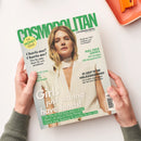 Schräg von oben fotografiertes, von zwei Händen mit grauen Strickärmeln gehaltenes Cosmopolitan-Magazin auf weißem Untergrund; das Cover zeigt eine blonde weibliche Person in weißem Oberteil sowie grüne und schwarze Texte wie „COSMOPOLITAN“, „FUN. FEARLESS. FEMALE.“, „THE MONEY ISSUE“, „Charis-ma? Charis-me!“ und „Girls just wanna have fonds!“. Oben rechts liegt ein orangefarbener Gegenstand.