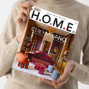Person in beigefarbenem Strickpullover hält das Magazin „H.O.M.E.“ (Ausgabe Juni–August 2023) vor neutralem Hintergrund, Titelzeile „WHERE DESIGN BECOMES H.O.M.E.“; darunter Hauptüberschrift „CIAO MILANO – Das Beste vom Salone del Mobile und der Milan Design Week“. Titelbild zeigt ein Wohnzimmer mit roten Sofas, farbigen Hockern, rundem Tisch und goldbraunen Wänden. Weitere Texte: „Neue Möbel“, „EuroCucina“, „Yacht-Special“.