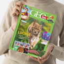 Helle Person in beigeem Pullover hält das Kindermagazin „GEOlino“ mit grünem Rand und Titelmotiv eines laufenden Luchses vor unscharfem Waldhintergrund. Auf dem Cover stehen u. a. die Texte „Luchse – Zurück in unseren Wäldern“, „Checker Tobi“, „Vulkane – Wenn die Erde Feuer spuckt“ und „Auf der Balz“. Unten links ist ein Foto von drei lachenden Kindern mit Berglandschaft im Hintergrund sichtbar.