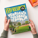 Magazincover „11 Freunde“ in gelber Schrift auf blauem Hintergrund, darunter große weiße Aufschrift „Verrückte Fußballrepublik“. Im unteren Bereich eine Park- oder Sportplatzszene mit mehreren Personen auf grünem Rasen und rotem Spielfeld. Oben rechts schwarzer Kreis mit gelber Schrift „Spieler der Saison – Die Wahl“. Hände halten das Magazin über eine helle Tischfläche neben orangefarbenem Behälter.