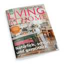 Titelbild der Zeitschrift „LIVING AT HOME“ mit großem pinkem Schriftzug auf hellem Hintergrund, darüber der Text „Mein Zuhause voller Ideen“; mittig eine helle Küche mit grünem Tresen, Holzhockern, gelben Zitronen in einer Schale, Vase mit Blumen, weiter hinten Esstisch mit Holzstühlen auf Fischgrätparkett; Text-Highlights: „Köstliche Eintöpfe“, „Natürlich schön und gemütlich“, „Garten-Saisonstart“, „Alles, was wir lieben“.