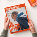 Zwei Hände halten ein schräg liegendes, orange-rot gerahmtes Magazin „DER SPIEGEL“ auf weißer Fläche. Das Cover zeigt eine männliche Person mit kurzem grauem Haar und Brille in dunkler Kleidung sowie blau gemustertem Stoff; lesbare Texte: „CSD-ANSCHLAG“, „GELDANLAGE“, „ZEN-KLOSTER“, „DIE PALAST-REVOLTE“ und „Union intern – Wie die eigene Partei an Merz verzweifelt“.