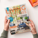Titelbild der Wohnzeitschrift „Living at Home“ mit orangefarbener Schrift und Unterzeile „Mein Zuhause voller Ideen“. Auf dem Cover ist ein modern eingerichtetes Wohnzimmer mit blauen und beigen Möbeln, großem Fenster und Pflanzen zu sehen. Weitere sichtbare Texte: „Einfach gut eingerichtet“, „Spargelrezepte“, „Per Rad durchs Ruhrgebiet“ sowie „Tolle Kombi Blau trifft Naturtöne“. Zwei Hände halten das Heft vor hellem Hintergrund.