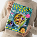Magazin „Slowly Veggie!“ wird von einer Person in beigefarbenem Strickpullover gehalten, Titel in gelber Schrift auf türkisfarbenem Hintergrund. Hauptüberschrift: „Rund um die Welt“, Unterzeilen u. a. „61 neue Lieblingsrezepte“, „Super Greens – schnelle Ideen mit Erbsen, Spinat & Kräutern“, „Urban Jungle“. Titelbild zeigt eine blaue Schale mit vegetarischem Gericht aus Gemüse, Quinoa und essbarer Blüte, daneben hellgelbe Sauce und grüne Essstäbchen.