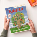 Titelbild der Zeitschrift „Servus Kinder“ mit Illustration eines großen Baums, auf dem neun Kinder spielen, lesen und klettern, umgeben von grüner Wiese, Zaun und Kuh im Hintergrund; Überschrift „Das Abenteuer deines Lebens“ sowie Text „Spielen | Staunen | Rätseln | Wissen“ und Kreis mit Aufschrift „Gewinne einen Urlaub am Bauernhof und vieles mehr“. Zwei Hände halten das Heft vor hell-beigem Hintergrund.