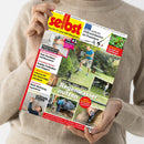 Helle, langärmlig gekleidete Person hält das Magazin „selbst ist der Mann – Das Do-it-yourself-Magazin“ in den Händen, Titelthema „Regenwasser nutzen“ mit Bildern von Personen bei Gartenarbeiten, grüner Rasen und Gartengeräten im Hintergrund; weitere sichtbare Schlagzeilen u. a. zu Elektroinstallation, Holz-Hochbeet, Fußballtor zum Klappen, Motorsägen-Schnitzen und Multi-Werkzeug-Reparatur.
