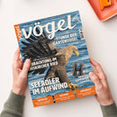 Titelseite des Magazins „vögel“ Nummer 87, Ausgabe 5/2025, gehalten von zwei Händen über hellem Tisch. Im Mittelpunkt ein fliegender Seeadler über blauem Wasser, rechts oben eine Abbildung eines Fernglases „ZEISS SFL 10x30“. Hauptüberschriften: „Stunde der Gartenvögel“, „Beobachtung im Eriskircher Ried“, „Seeadler im Aufwind“. Untere Leiste in Orange mit Themen: Erforschung, Bestimmung, Aufgedeckt.