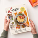 Titelblatt der Zeitschrift „Servus – einfach gut leben“ mit großem Foto einer Dessertschale mit Vanilleeis und Erdbeeren auf weißem Teller mit Dekogras. Kreisförmiger gelber Hinweis „Wie werde ich glücklich?“ rechts oben, darunter Überschrift „Oh, süßer Mai!“. Links kleinere Fotos mit den Themen „Leben in Dreieichhof“ und „Heimat im Quartal“. Gehalten von zwei Händen vor hellbeigem Hintergrund.