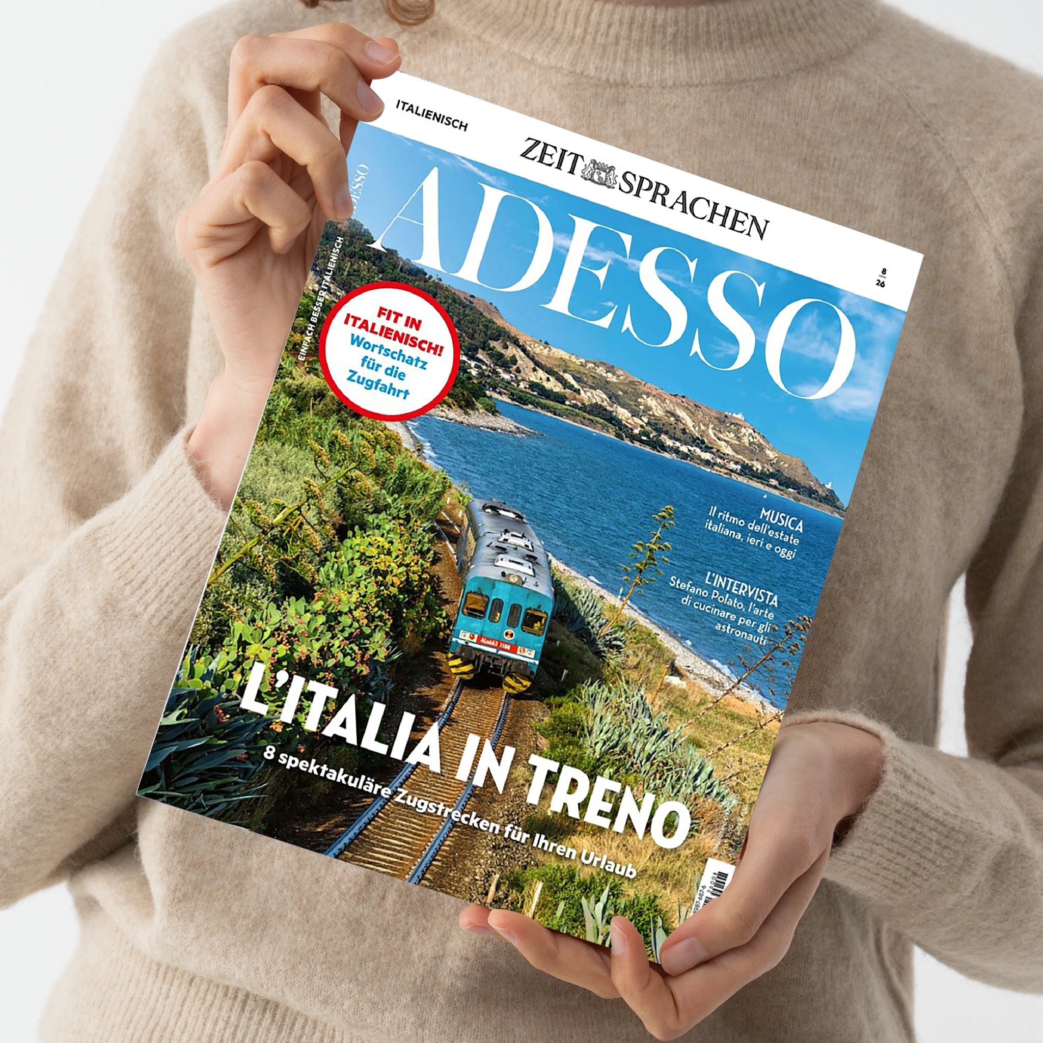 Person in beigefarbenem Wollpullover hält die Zeitschrift „ADESSO“ aus der Reihe „ZEIT SPRACHEN“ mit blauem Titelkopf und großem Foto eines blauen Zuges auf Küstenstrecke in Italien, flankiert von grüner Vegetation und Meer. Sichtbare Texte: „L’ITALIA IN TRENO“, „8 spektakuläre Zugstrecken für Ihren Urlaub“, roter Kreis mit „FIT IN ITALIENISCH – Wortschatz für die Zugfahrt“, sowie Rubriken „MUSICA“ und „L’INTERVISTA“.
