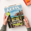 Das Magazin „ECOS“ von „ZEIT SPRACHEN“ liegt schräg auf einem weißen Tisch und wird von zwei Händen mit grauen Strickärmeln gehalten. Das Cover zeigt eine Küstenlandschaft mit blauem Meer, Felsen und grünen Kakteen; lesbare Texte sind „SPANISCH“, „GRACIELA ITURBIDE“, „WORTSCHATZ Vacaciones en el Caribe“, „Baja California“ und „Roadtrip in Mexiko von Tijuana bis Los Cabos“.