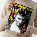 Person in beigefarbenem Pullover hält ein Magazin mit schwarzem Cover und großem Schwarz-Weiß-Porträt eines jungen Mannes mit welligem Haar. Gelber Schriftzug „musikexpress“ oben, weitere Texte in Gelb und Pink: „40 Jahre ‘Hunting High and Low’“, „A-HA – Alles über ihr Meisterwerk“, „Die 25 besten unbekannten Pop-Alben der 80er“, „Lana Del Rey – Kaliforniens dunkle Königin“, „Pop gegen Rechts – Im Osten regt sich Widerstand“.