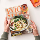 Titelblatt der Zeitschrift „Food and Travel Deutsch“, gehalten von zwei Händen über hellem Tisch. Großes orangefarbenes Titelwort „FOOD and TRAVEL“; weitere Aufschriften: „Rezepte für den Frühling“, „Neue Safari-Lodges“, „Stadthotels unter 200 €“, „Malaysias Gourmetinsel Langkawi“, „Kulinarik-Hotspot Cadiz“, „Skifahren im Spätwinter“, „Argentinien Córdoba“. Im Hintergrund eine Schüssel Nudelsuppe mit Gemüse und Stäbchen, warme Farbtöne, matte Papieroberfläche.