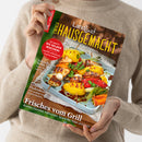 Weibliche Person in beigefarbenem Strickpullover hält ein schräg ausgerichtetes, glänzendes Magazin mit dem Titel „Landlust HAUSGEMACHT“ und dem Untertitel „Mein Magazin für Haus und Küche“. Sichtbare Texte: „So geht’s: Eis selber machen!“, „Schnelles für heiße Tage“, „Marmeladen“, „Gegrillte Fladenbrote“ und „Frisches vom Grill“; darunter ein Foto mit gegrilltem Gemüse und Grillspießen.