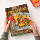 Schräg gehaltenes, grün gerahmtes Magazin mit dem Titel „Landlust EIN HAUSGEMACHT!“ und dem Untertitel „Mein Magazin für Haus und Küche“. Das Cover zeigt gegrilltes Gemüse und Fleisch; lesbare Texte sind „Marmeladen“, „Eis selber machen!“, „Frisches vom Grill“ sowie „Buntes Gemüse, Hähnchen, Spareribs, Käse und Fisch“; zwei Hände mit grauen Ärmeln halten das Magazin, eine Person ist nicht erkennbar.