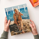 Schräg auf einer weißen Fläche liegendes, von zwei Händen einer nicht erkennbaren Person gehaltenes ELLE-Magazin mit glänzendem Papier und einer blonden Frau im Muscheloberteil am Strand vor blauem Meer. Sichtbare Texte: „ELLE“, „LOVE IT! 9 Schritte zu richtig gutem Sex“, „BIG SPLASH Die besten Hitze-Hacks“, „DAS SONNEN-HOROSKOP“ und „summer in STYLE“.