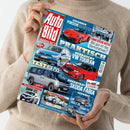 Person in beigem Strickpullover hält eine Zeitschrift mit dem Titel „Auto Bild“ in rotem Logo. Auf dem Cover sind mehrere Autos in verschiedenen Perspektiven abgebildet: oben ein roter Porsche 911 GT3 S/C, mittig ein silberner VW Touran und ein roter Skoda Fabia, unten ein silberner Kia PV5, BMW iX3, Audi Lucca, Geely E5 und Zeekr 7X. Schlagzeilen u. a. „Praktisch – gebraucht VW Touran“, „Test Kia PV5“, „Gefahren Porsche 911 GT3 S/C“, „Chinesische Autos“.