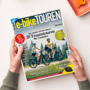 Titelbild des „e-bike TOUREN MAGAZIN“ mit der Aufschrift „20 Traumtouren für Genießer“, „Märchenhafte Mittelgebirge“ und „Marktcheck E-Bike-Motoren“. Abgebildet sind eine weibliche und eine männliche Person mit Helmen auf E-Bikes auf einem Waldweg, dahinter grüne Tannen und blauer Himmel. Das Magazin wird von zwei Händen mit grünem Pullover vor hellem Hintergrund gehalten.