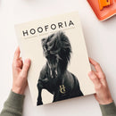 Helle Zeitschrift mit dem Titel „HOOFORIA“ und dem Untertitel „NACHDENKEN • BEWEGEN • VERSTEHEN • BEGEISTERN“, mittig ein großes schwarz-weiß-Foto eines laufenden Pferdes mit wehendem Schweif, unten das goldfarbene Logo „H C VOLUME I“, gehalten von zwei Händen in grünen Strickärmeln, auf hellem Hintergrund, oben rechts teilweise sichtbare orangefarbene Schale.