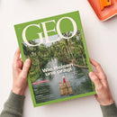 Titelseite des GEO-Magazins, Ausgabe Juni 2023, mit großem weißen Schriftzug „GEO“ auf grünem Hintergrund. Darunter farbfotografische Aufnahme eines tropischen Flusses mit dichter Palmenkulisse; im Vordergrund steht eine einzelne Person in rotem Gewand auf einem Bambusfloß. Hauptüberschrift: „Wie Reisen uns prägt“, weitere Texte zu Hirnforschung, Mali und Atommüll. Zwei Hände halten die Zeitschrift über hellem Tisch.