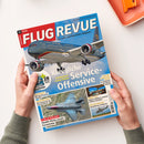 Von zwei Händen einer nicht näher bestimmbaren Person gehaltenes, schräg liegendes Magazin „FLUGREVUE“ auf weißem Untergrund. Das Cover zeigt ein Passagierflugzeug vor blauem Himmel sowie kleinere Flugzeug- und Hubschrauberbilder; lesbare Texte sind „Das Luft- und Raumfahrt-Magazin“, „Königliche Service-Offensive“, „Jordaniens neue Flotte“, „F-35 in Italien“ und „Boeing 777-9“.