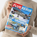 Eine Person hält eine schräg fotografierte Zeitschrift auf einem grob gestrickten beigefarbenen Pullover. Das rot-blaue Cover trägt den Titel „FLUG REVUE – Das Luft- und Raumfahrt-Magazin“ und zeigt ein Passagierflugzeug; sichtbare Texte sind „Königliche Service-Offensive“, „Jordaniens neue Flotte“, „F-35 in Italien“, „Helikopter“ und „28 Seiten extra“.