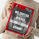 Person in beigefarbenem, grob gestricktem Pullover hält eine schräg fotografierte Zeitschrift mit rotem Rand und schwarzem Titelbild in beiden Händen. Sichtbare Texte: „FOCUS“, „NEUROZWEIFELN WIRD DER ISLAMIS... GEFAHR?“ sowie „Endspiel für den Kanzler“, „Wirtschaft unter Schock“ und „Nach dem Anschlag von Berlin: Der Staat darf sich nicht länger vorführen lassen.“