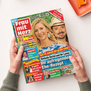 Aufrecht gehaltene Zeitschrift „Frau mit Herz“ mit rotem Cover, fotografiertem blondem weiblichem Model und männlichem Model mit Schnurrbart und Strohhut. Sichtbare Texte sind „Extra großer Rätselspaß“, „Beatrice Egli: Baby-Sehnsucht“, „Süße Pläne fürs Familienglück“, „Seit 15 Jahren die große Liebe“ und „Stefanie Hertel und ihr Lanny: Ihr aufregendes Ehe-Rezept“; zwei Hände halten das Magazin.