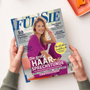 Schräg gehaltene Zeitschrift „FÜR SIE“ mit einer lächelnden blonden Frau in pinker Kleidung auf dem Cover, umgeben von zwei Händen in grauen Strickärmeln. Sichtbare Texte: „50 Sommer-Highlights“, „Stress lass nach!“, „Die Liebe auffrischen“, „Die große Haar-Sprechstunde“ und „Wunderbare Landküche“; im Hintergrund liegt teilweise ein orangefarbenes Tablett.