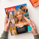 Hochformatige, glänzende „Gala“-Zeitschrift auf weißer Fläche, schräg von oben fotografiert und von zwei Händen mit graugrünen Ärmeln gehalten; das Cover ist rot, blau, türkis und orange gestaltet und zeigt eine blonde Frau sowie kleinere Fotos mehrerer Frauen und Männer. Lesbare Texte: „Traut sie sich?“, „Jennifer Aniston“, „Victoria & David Beckham – Endlich Frieden?“, „Phil Collins – Sein Sohn kickt in Bayern“, „Leah Isadora“ und „Ariana Grande – Große Sorge“.