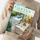 Person in beigefarbenem Strickpullover hält ein türkis eingefasstes Garten- und Wohnmagazin. Das Cover trägt den Titel „GARTEN WOHNEN“ und zeigt eine Gartenanlage mit Pool, Terrasse und gedecktem Esstisch; sichtbare Texte: „Genießen wie im sonnigen Süden“, „Cloud Dancer“, „Côte d’Azur“ und „Lavendelfeldern“.