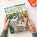 Hochformatige Zeitschrift „GARTEN & WOHNEN Träume“ mit türkisfarbenem Rand, gehalten von zwei Händen; das Cover zeigt einen Garten mit Pool sowie eine überdachte Terrasse mit gedecktem Esstisch. Lesbare Texte: „Genießen wie im sonnigen Süden“, „Cloud Dancer“, „Verliebt in die Provence“ und „Erfrischende Ideen von Brunnen bis Wasserbecken“.
